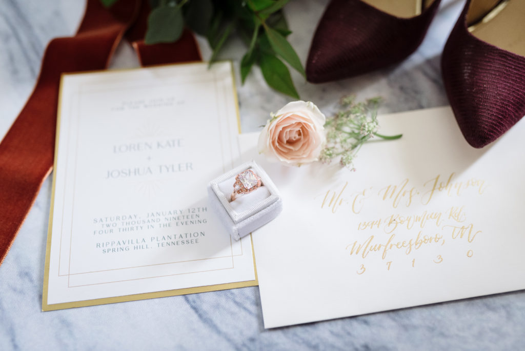detail shot of a wedding invitation suite with wedding shoes and wedding ring at a january wedding featuring lush red tones