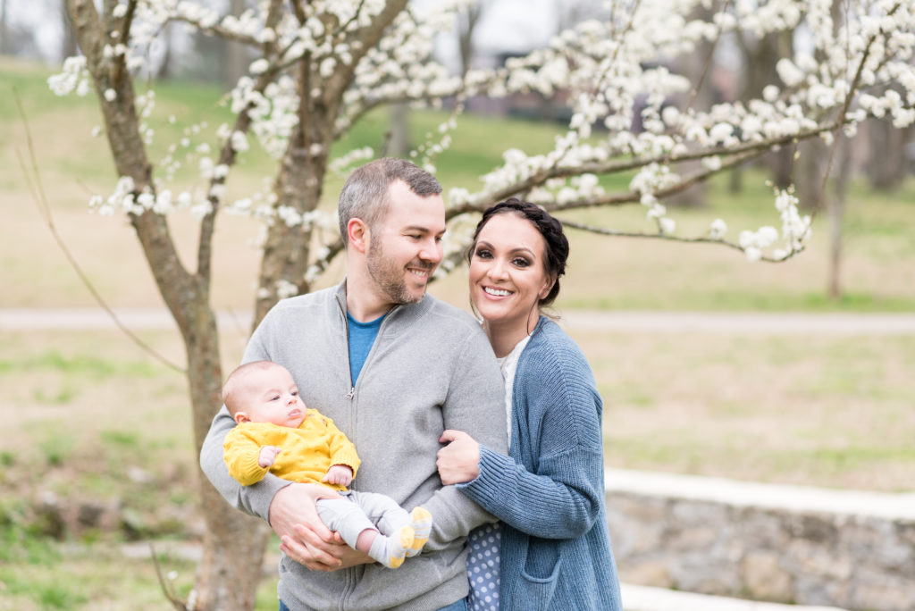 Family of three portraits under a cherry tree for Spring. Husband is looking at wife and smiling and wife is smiling at the camera while baby looks off because well, he's a baby!