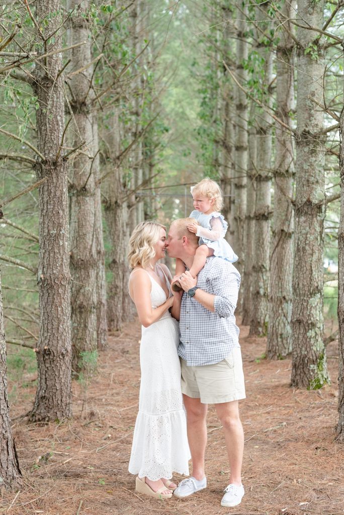 Mom and Dad are kissing and Dad at the same time is holding toddler aged girl on his shoulders as she watches her parents kiss in a forest