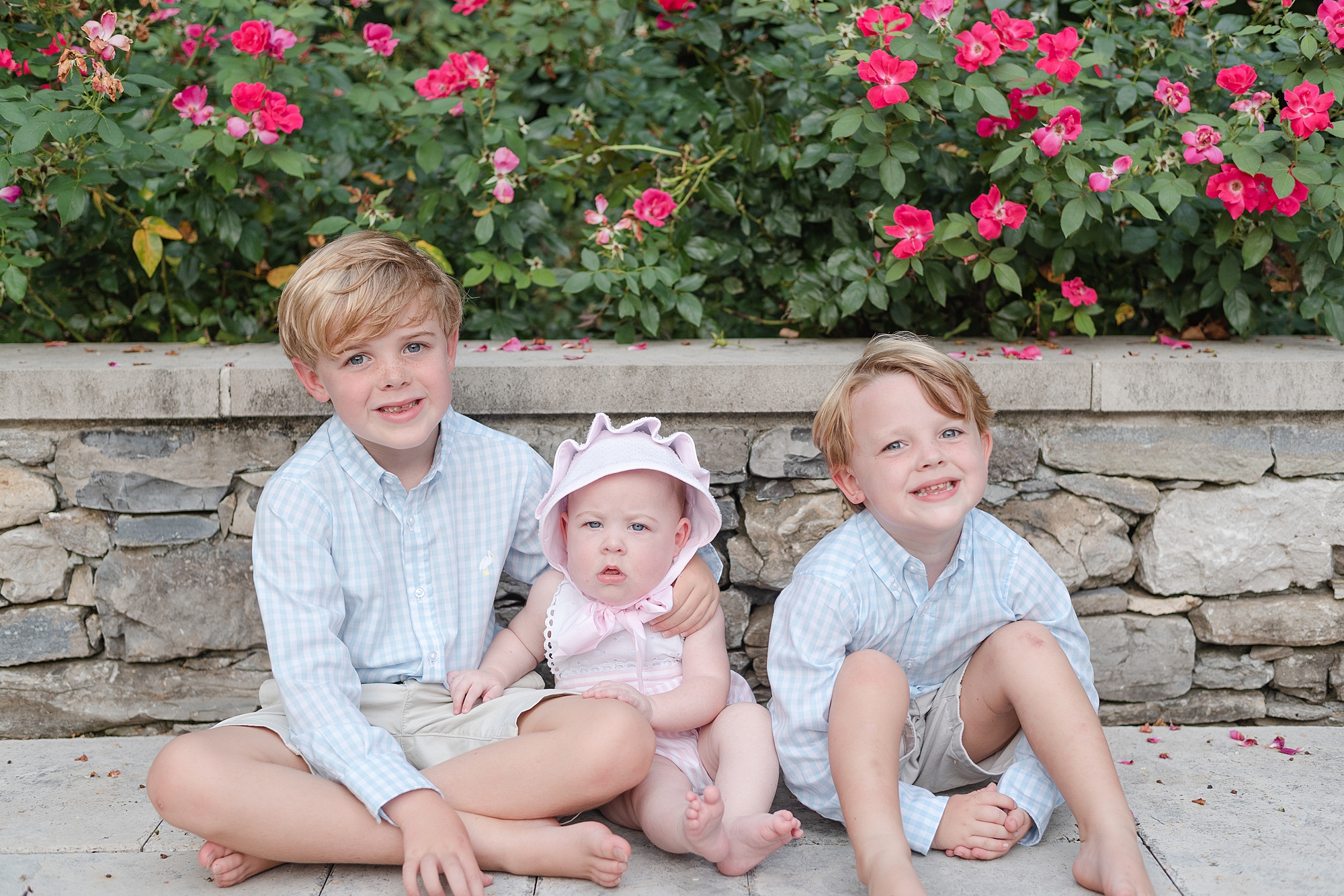 Two older brothers and baby sister are sitting along a stone brick wall with roses behind them and are smiling at the camera for a sibling photo