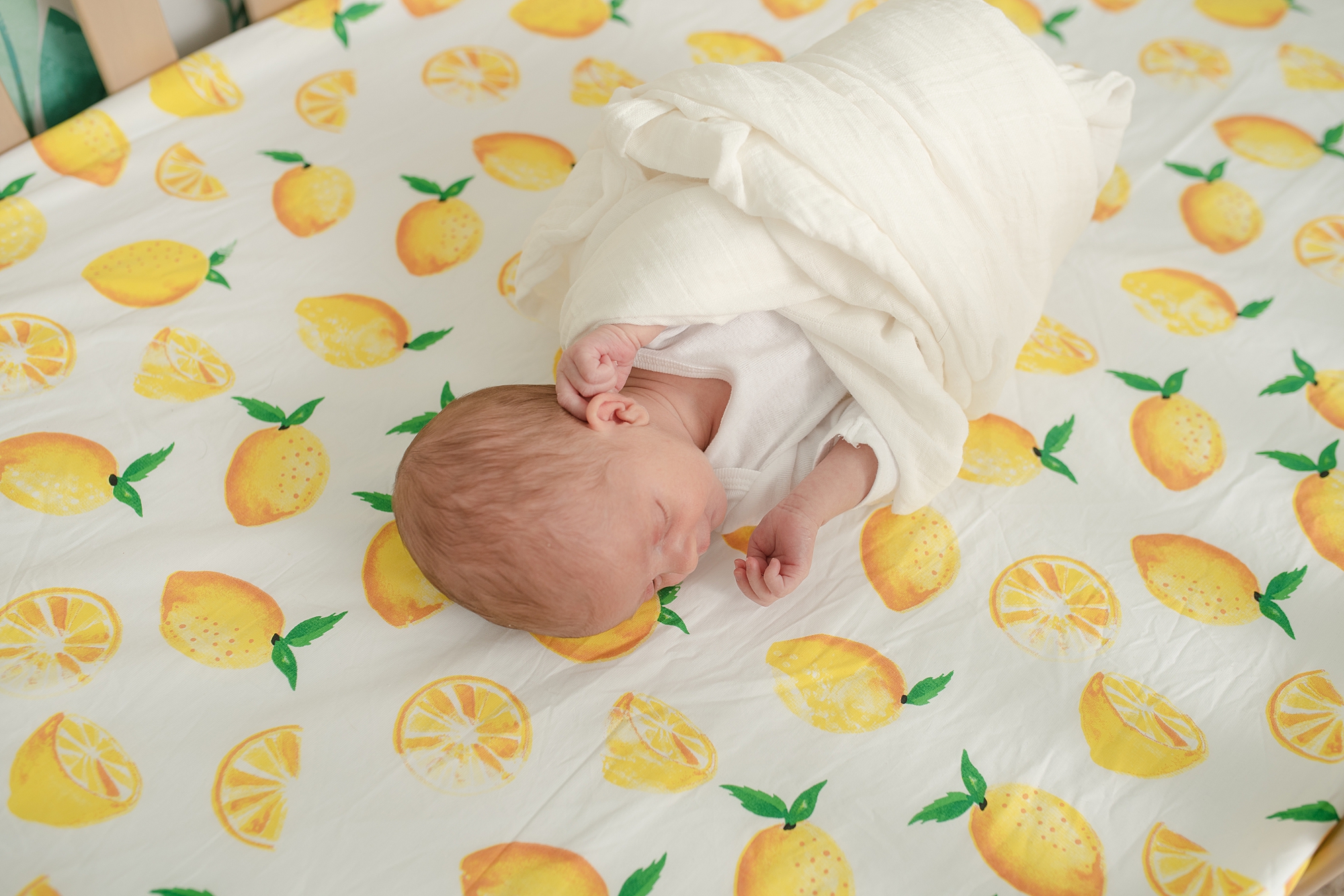 baby girl laying in her crib is swaddled in a white swaddle and is laying down on her cribsheets which are lemon print
