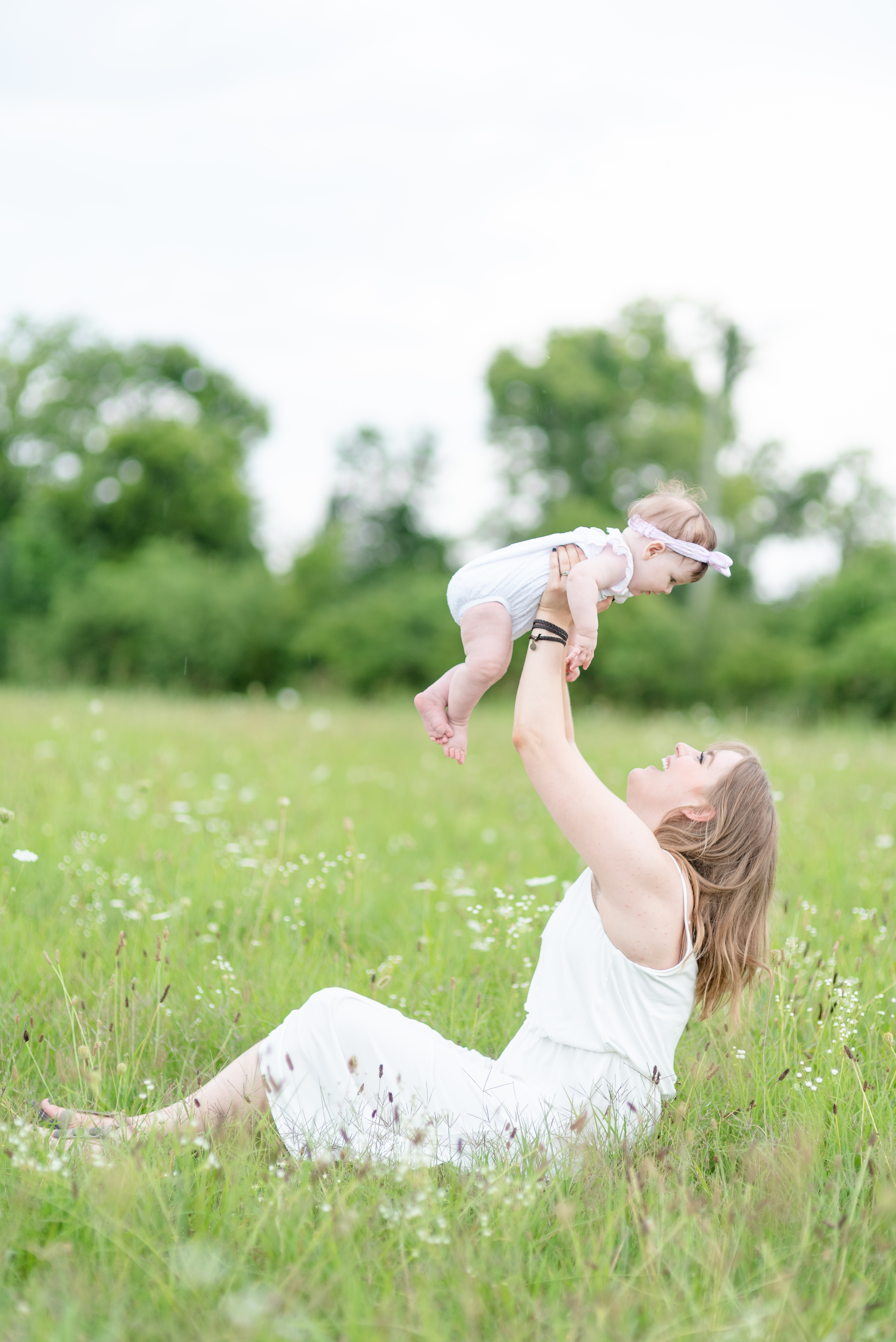 Mother Wearing A White Dress Holding her Baby Daughter in the Air while sitting in a field of flowers