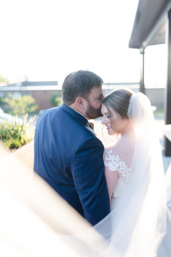 A husband kisses his new bride on her forehead in the soft glow of the golden hour light