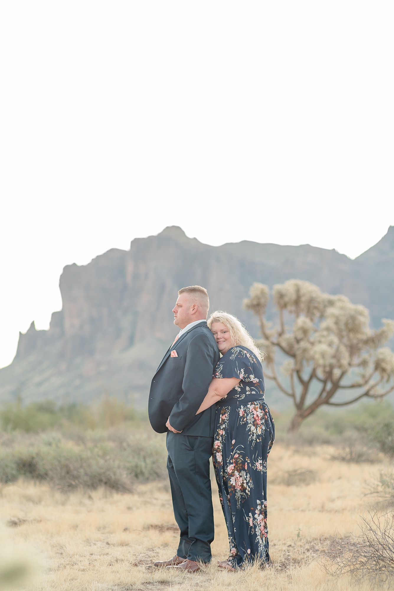 A Fiance holds her husband to be and he looks off camera and she looks at the camera for their Arizona Sunrise Engagement Session At The Paseo by Destination Wedding Photographer Dolly DeLong Photography