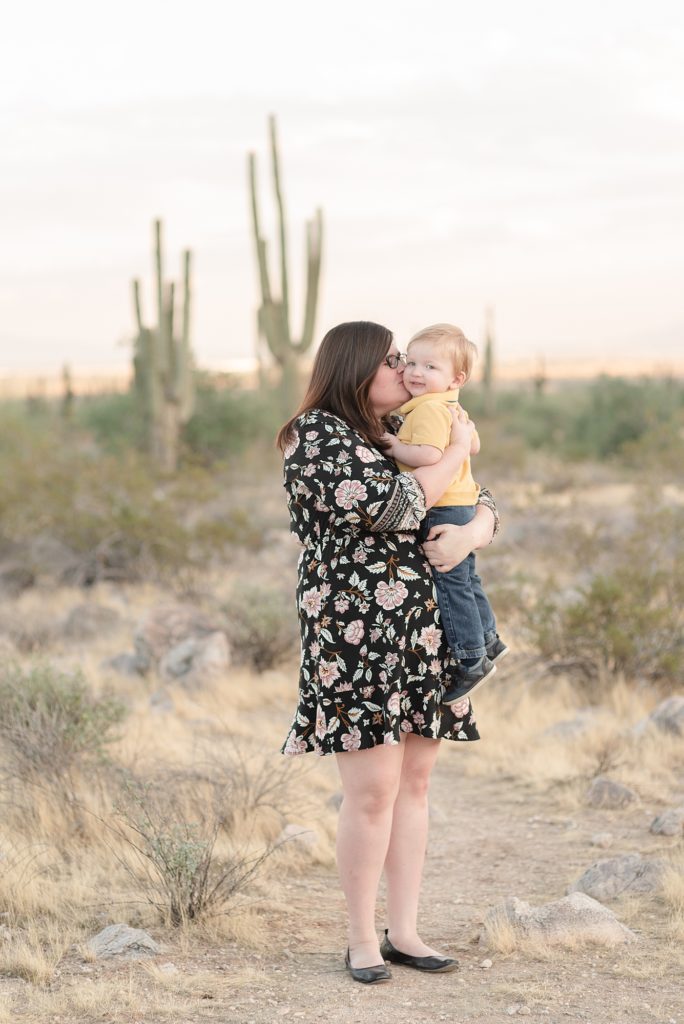A mom is snuggling her youngest son in her arms and he is looking at the camera and grinning in the Phoenix Arizona Desert Near the White Tank Mountains for christmas portraits