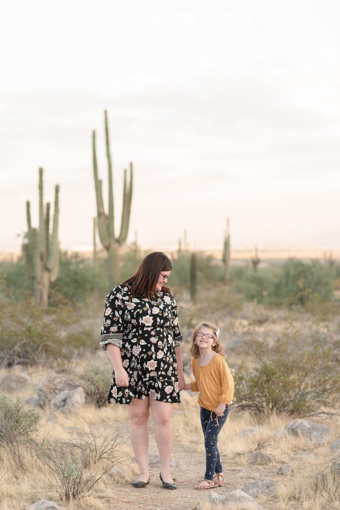 A mom is holding her oldest daughters hand and is looking down at her daughter and the daughter is smiling at the camera for family portraits in Phoenix Arizona at the base of the White Tank Mountains