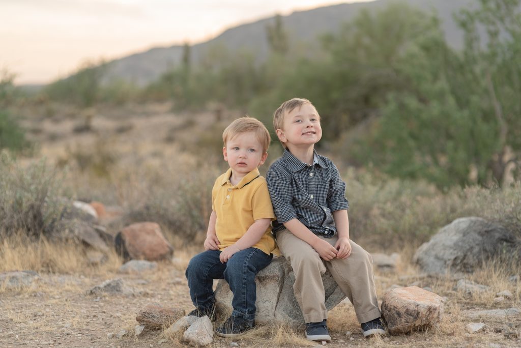 Two brothers are sitting on a rock and smiling for family portraits in the Phoenix Arizona Desert at the base of the White Tank Mountains