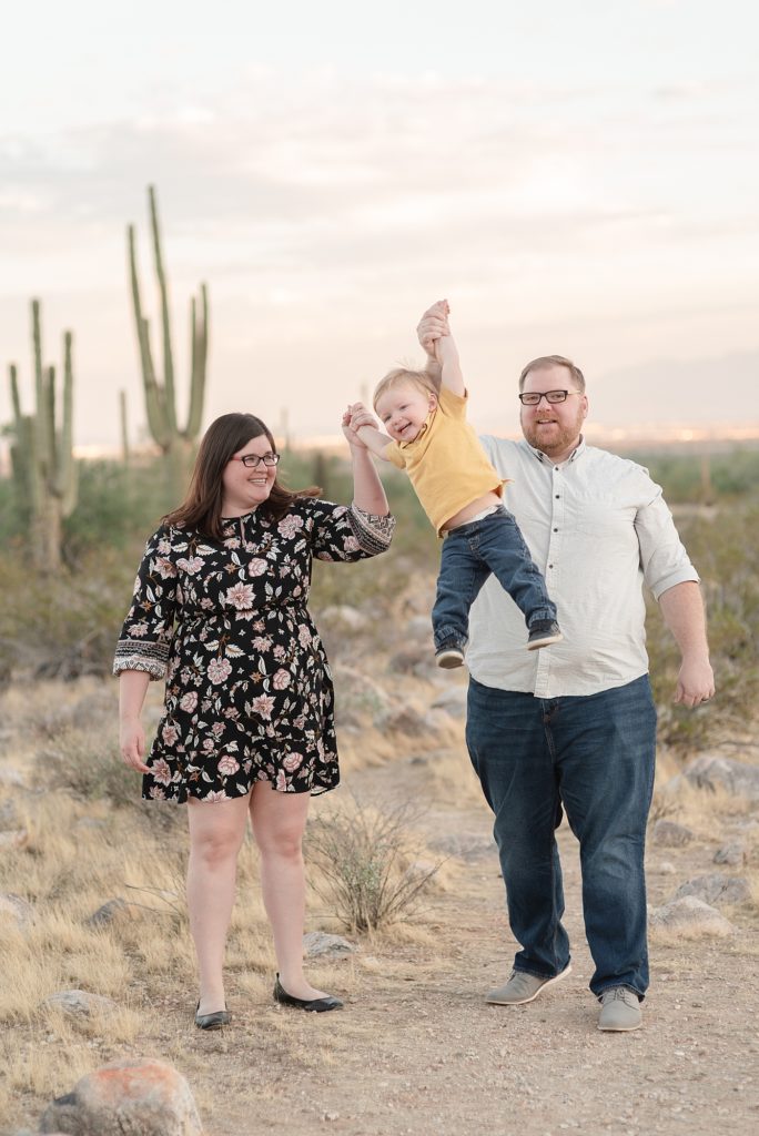 A mom and dad are swinging their toddler in the air for family portraits in the Phoenix Arizona Desert at the Base of the White Tank Mountains