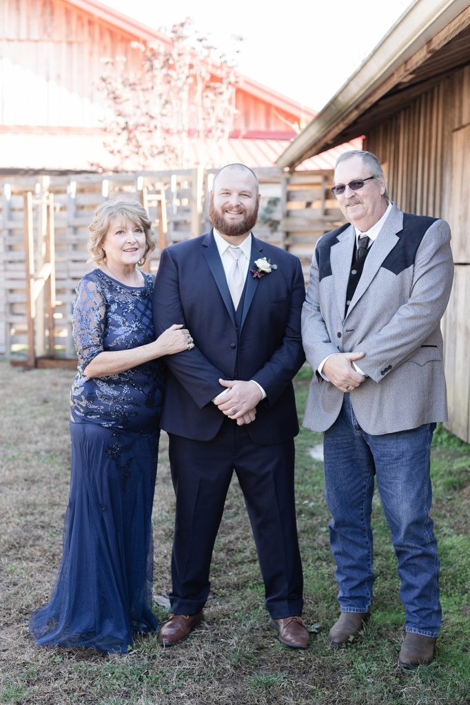 a groom standing by his mother and father on his wedding day while wearing a blue navy tuxedo and smiling