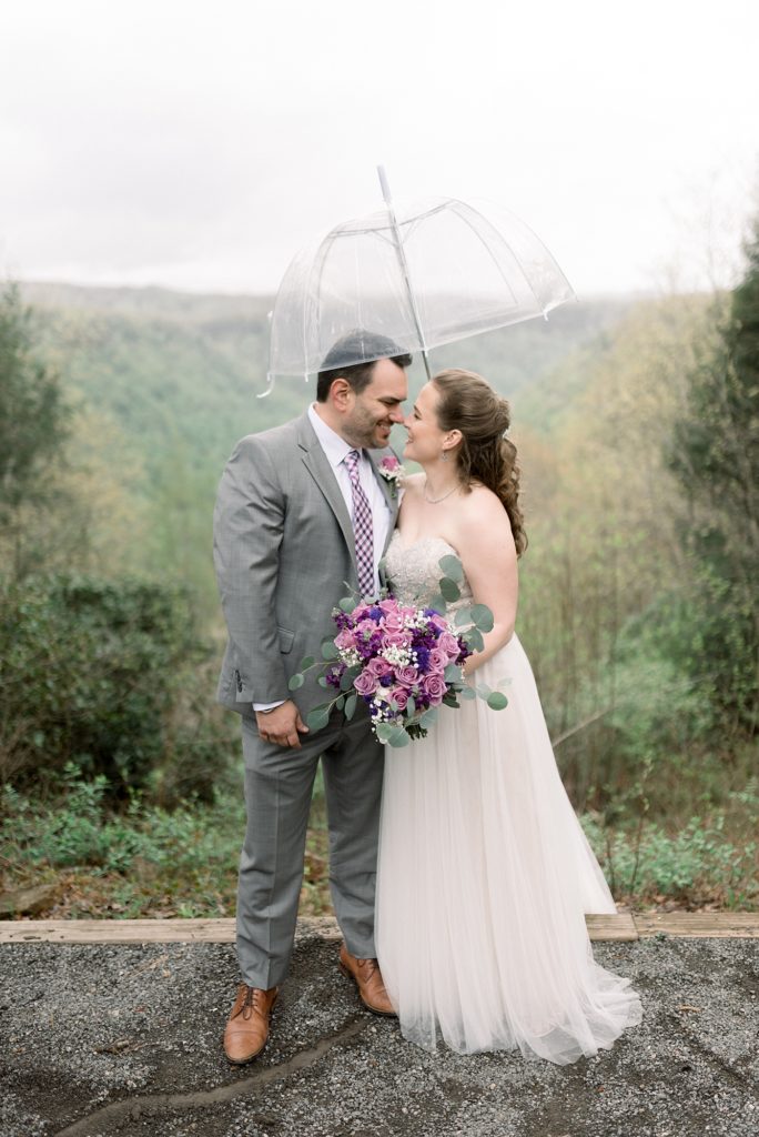 A bride and groom are smiling at eachother under a clear umbrella for their wedding at Adventures On The Gorge by Dolly DeLong Photography