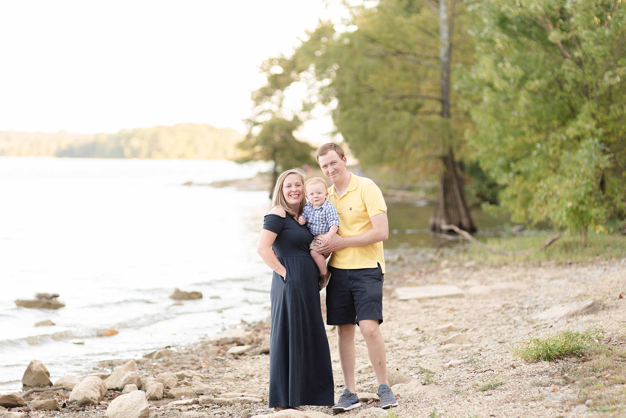 family of three standing beside lake and smiling at camera for family portraits by DollyDeLong