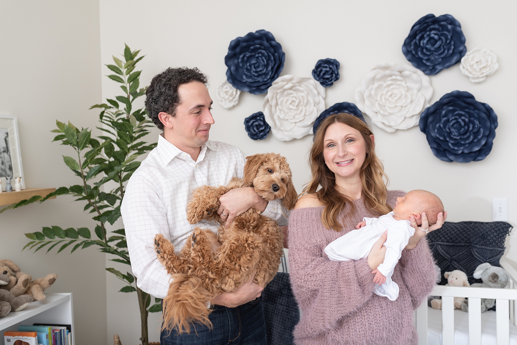 a dad is holding a dog and a mom is holding her newborn daughter in the daughter's nursery in Nashville for a funny family photo by Dolly DeLong Photography