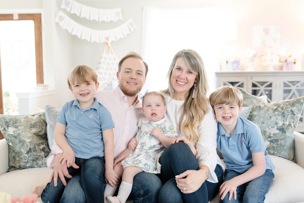 a family of five are sitting on a white couch and smiling at the camera for their little sister's first birthday party in Nashville by Nashville family photographer Dolly DeLong Photography