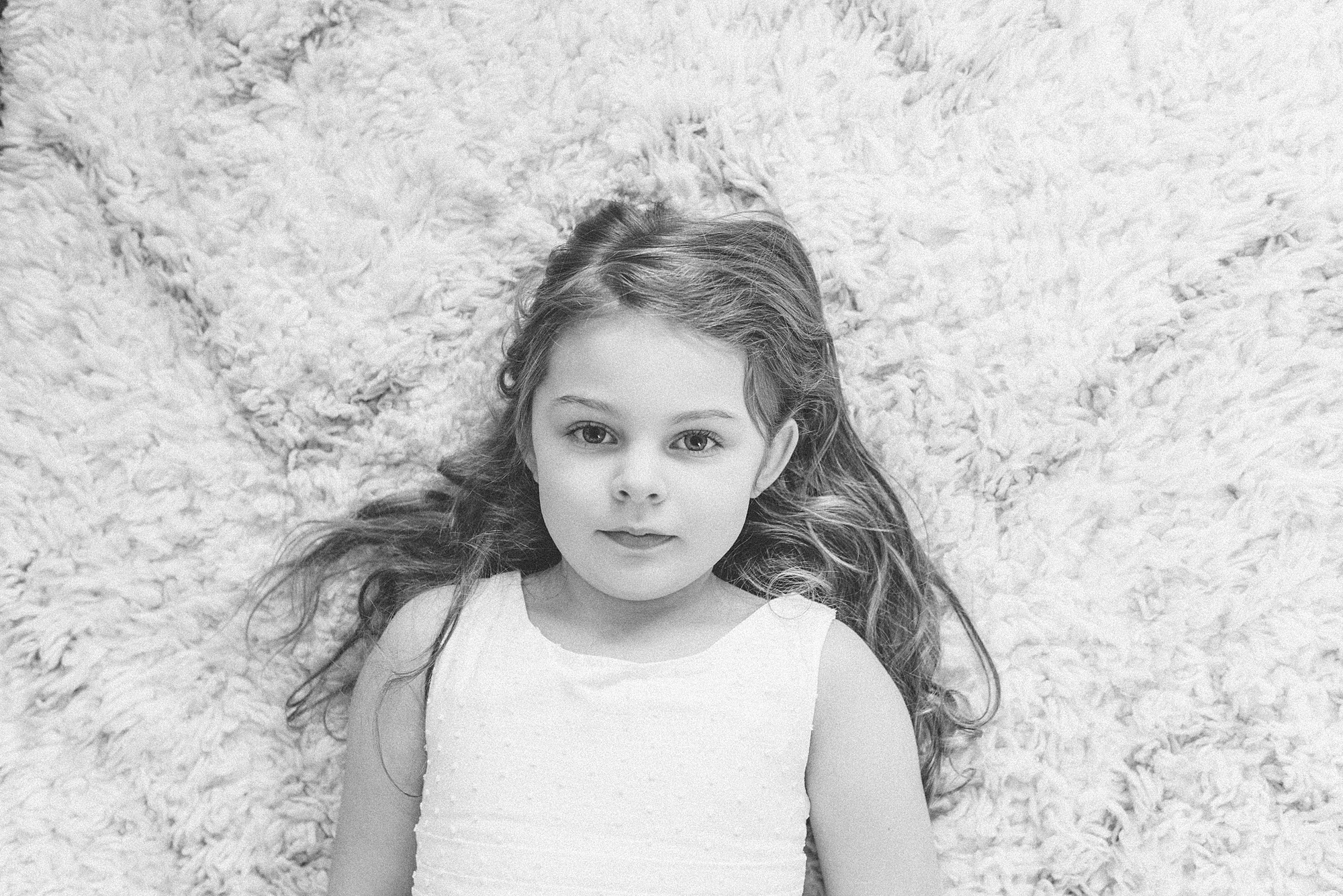 a black and white photo of a little girl laying on her back on a rug and looking up at at the photographer by nashville family photographer dolly delong photography