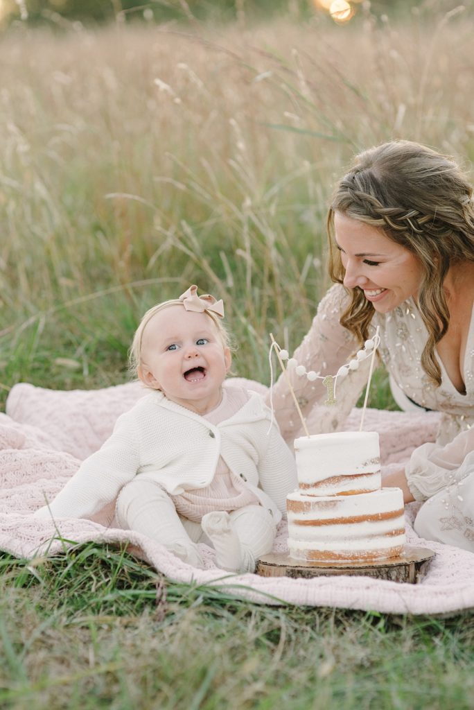 a one year old sitting on a blanket in a tall grassy field for her milestone photos with cake by Nashville Family Photographer Dolly DeLong Photography