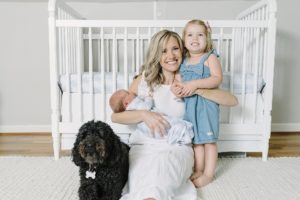 mom sits in front of crib with daughter, son, and dog