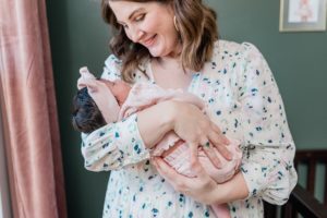 mom looks down at baby girl in arms standing by window