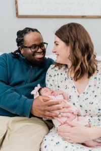 mom and dad smile holding baby girl on bed during newborn photos at home