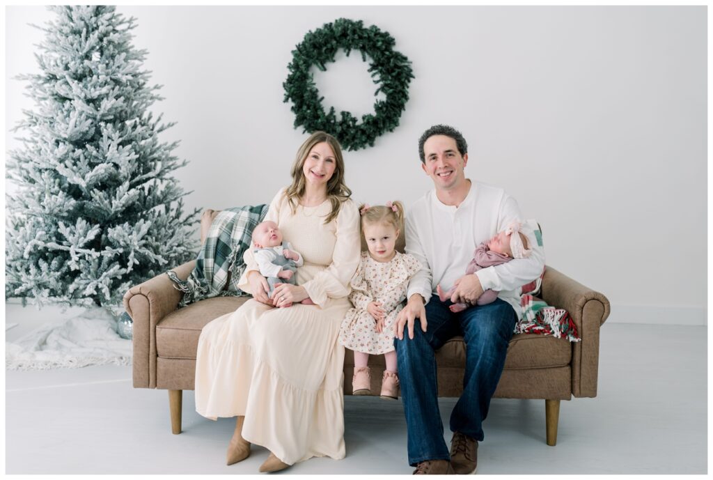 Mom_and_Dad_are_holding_their_twins_with_their_older_daughter_sitting_In_the_middle_on_a_tan_couch_for_holiday_photos_in_Vow_Collective_Studios