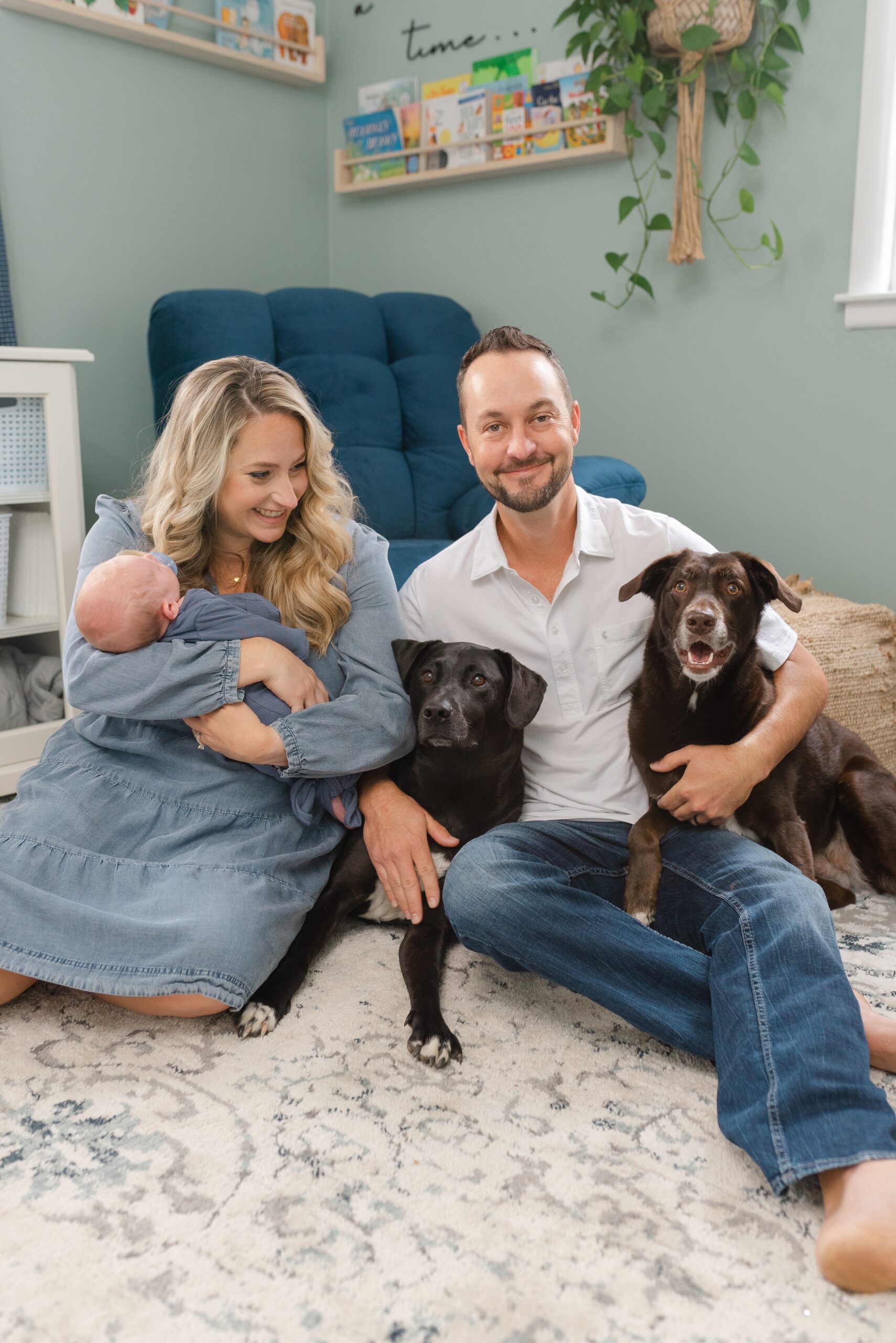 a_mom_and_dad_smile_at_at_camera_while_holding_their_newborn_son_and_their_pet_dogs_at_their_nashville_newborn_photography_Session_With_Dolly_DeLong_Photography