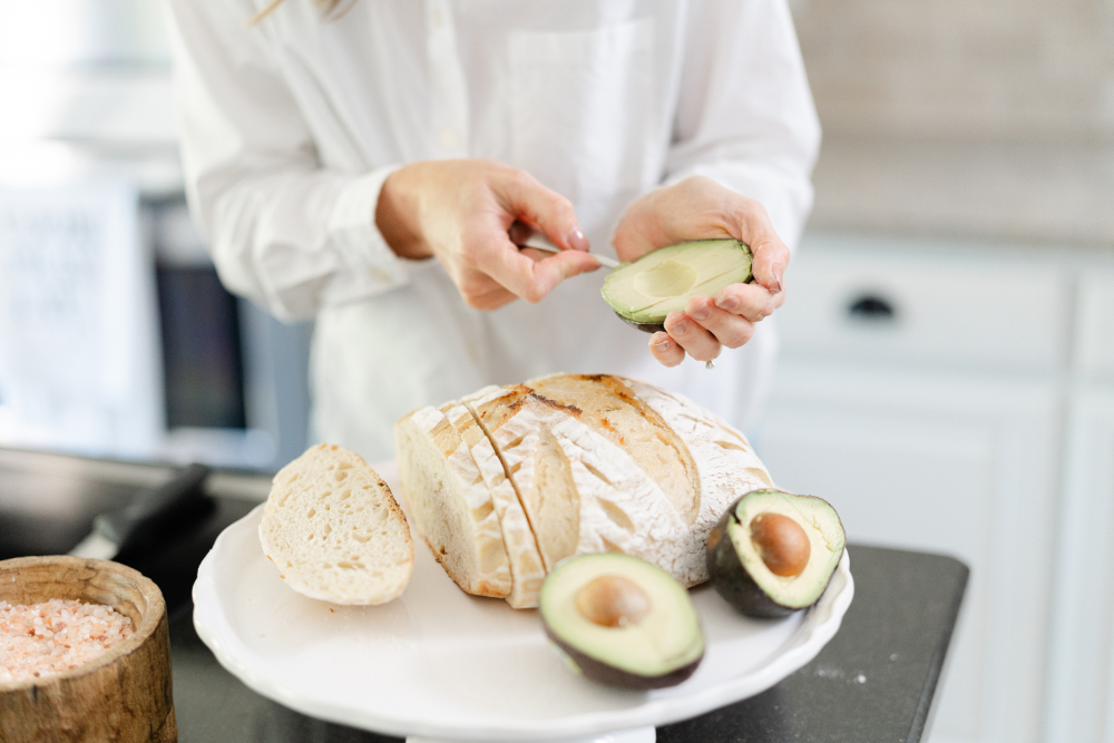 Close-up of hands scooping an avocado next to sliced bread and additional avocado halves on a plate.