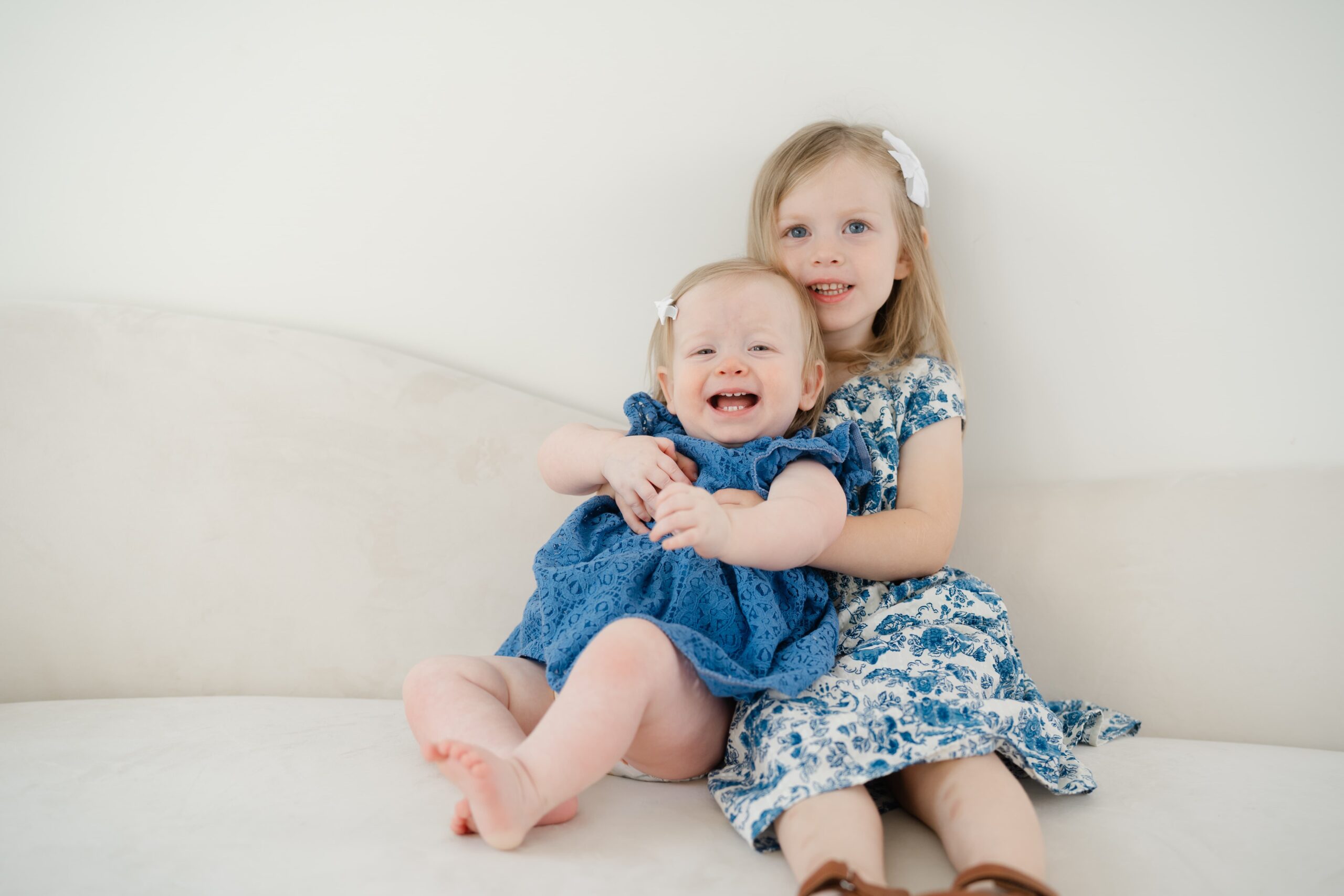 big-sister-holding-baby-sister-and-smiling-on-a-white-couch-at-studio-ivoire