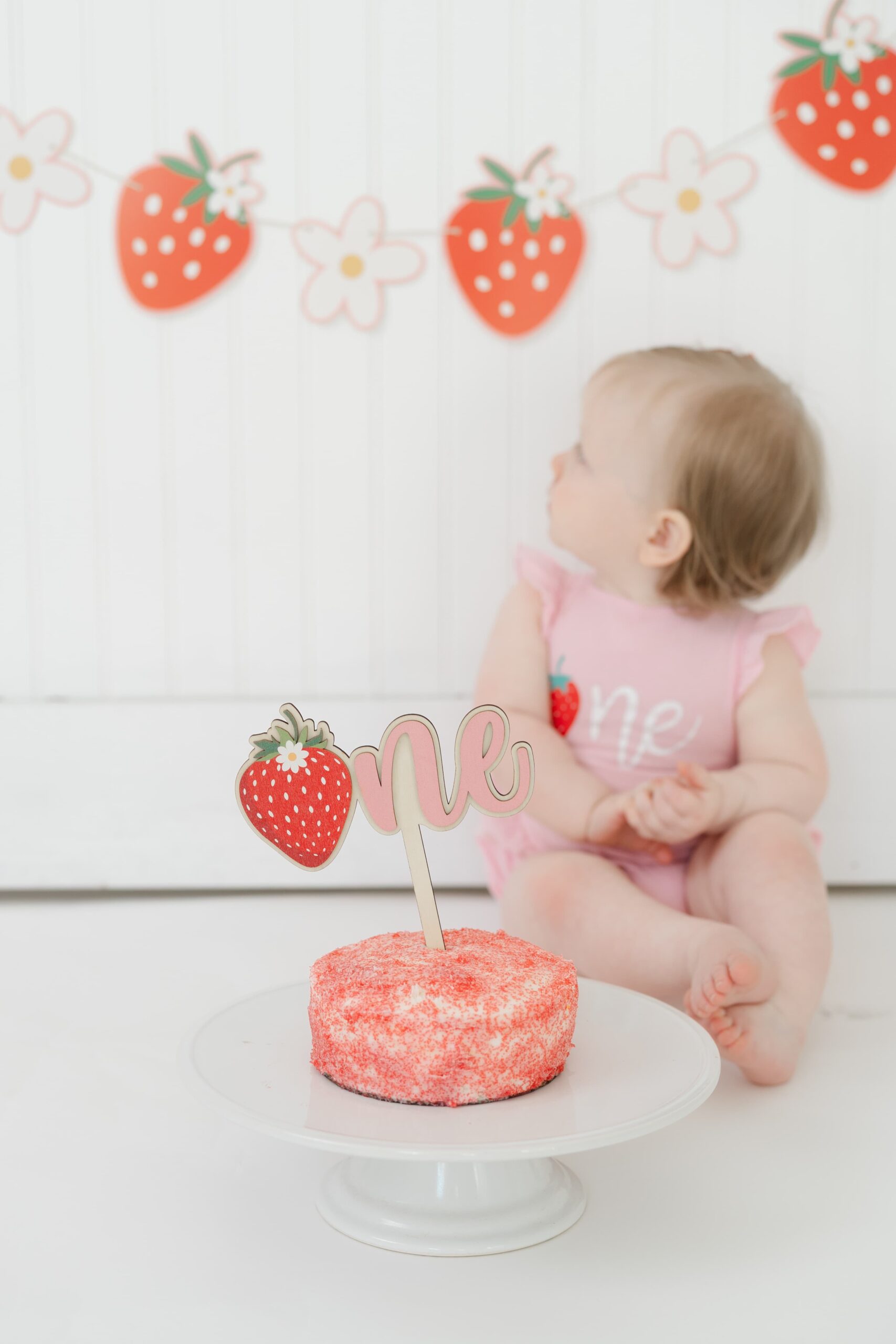 baby-girl-looking-at-strawberry-banner-with-cake-in-foreground-during-cakesmash-session-in-franklin-tennessee