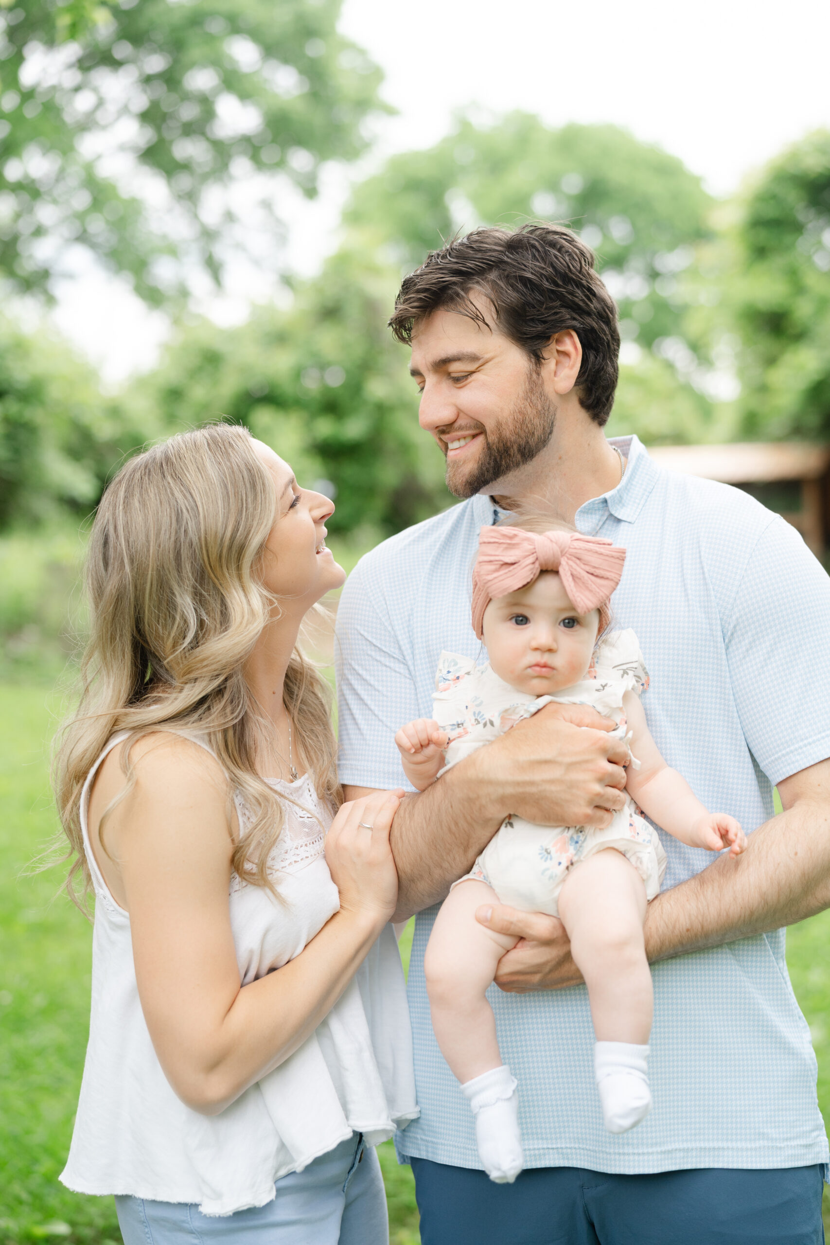 Parents smiling lovingly at each other while holding their baby girl outdoors, capturing a sweet candid family moment.