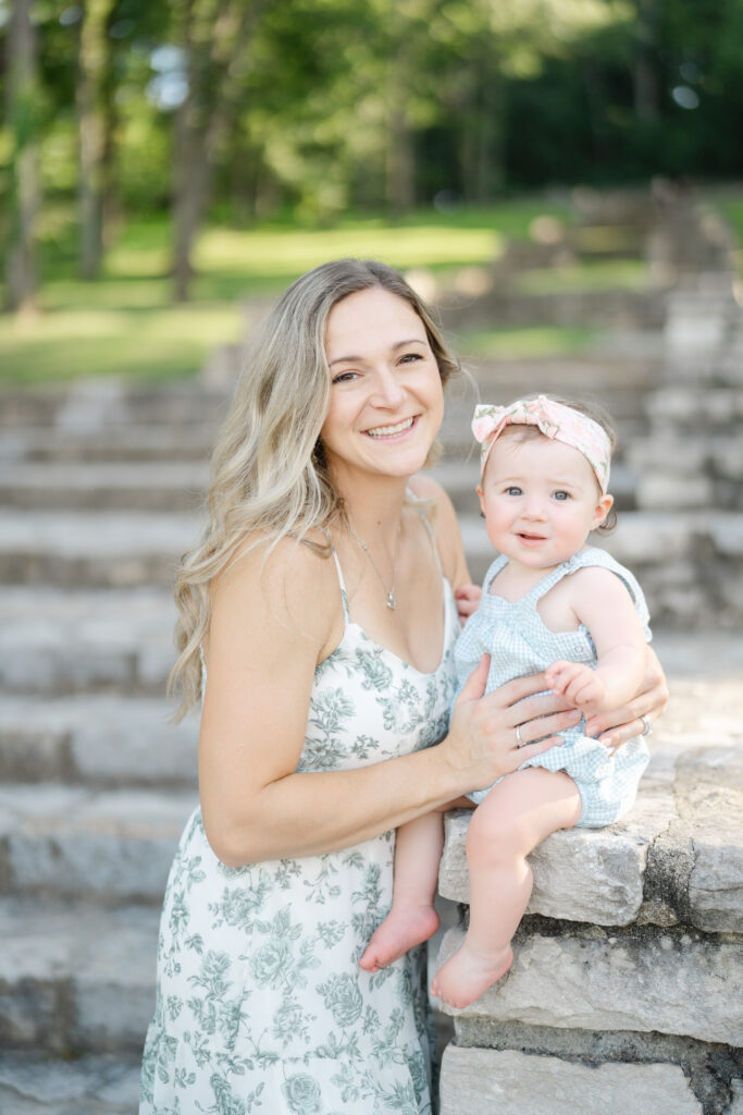 Mother holding her baby girl with a large pink bow during a bright outdoor milestone photo session.