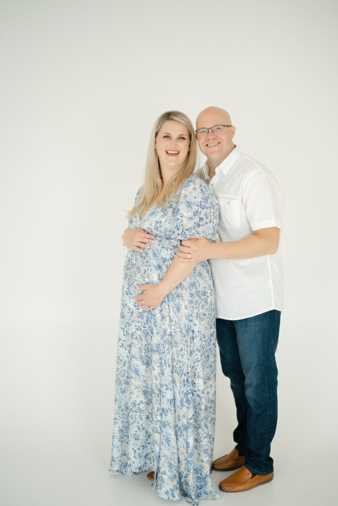 Expecting parents posing in a light blue floral dress and casual white shirt — timeless example of what to wear for maternity photos in a studio setting.