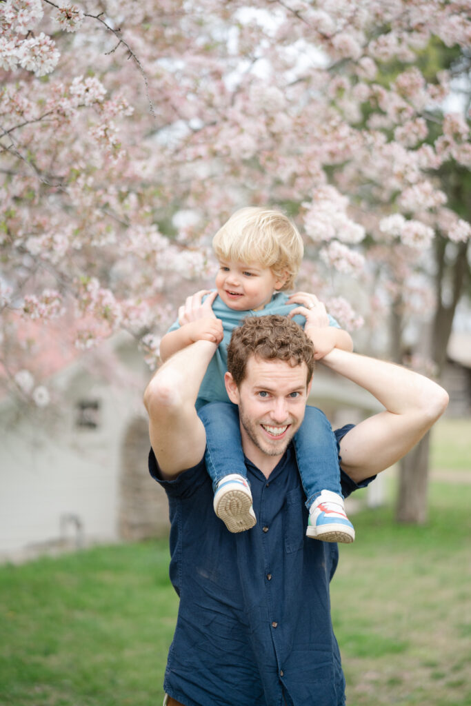 Dad giving toddler a shoulder ride under blooming cherry blossoms, both smiling during a playful spring session. family photography poses