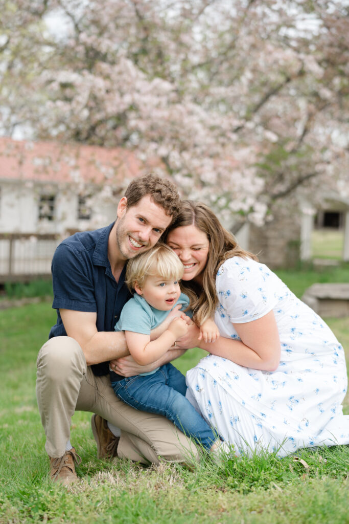 Family kneeling together in front of flowering trees, hugging their toddler for a sweet candid moment, a gentle family photography poses idea.