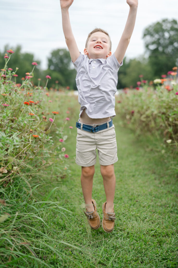 Young boy jumping in a flower field with arms raised, joyful energy and movement captured during a playful family session. family photography poses.