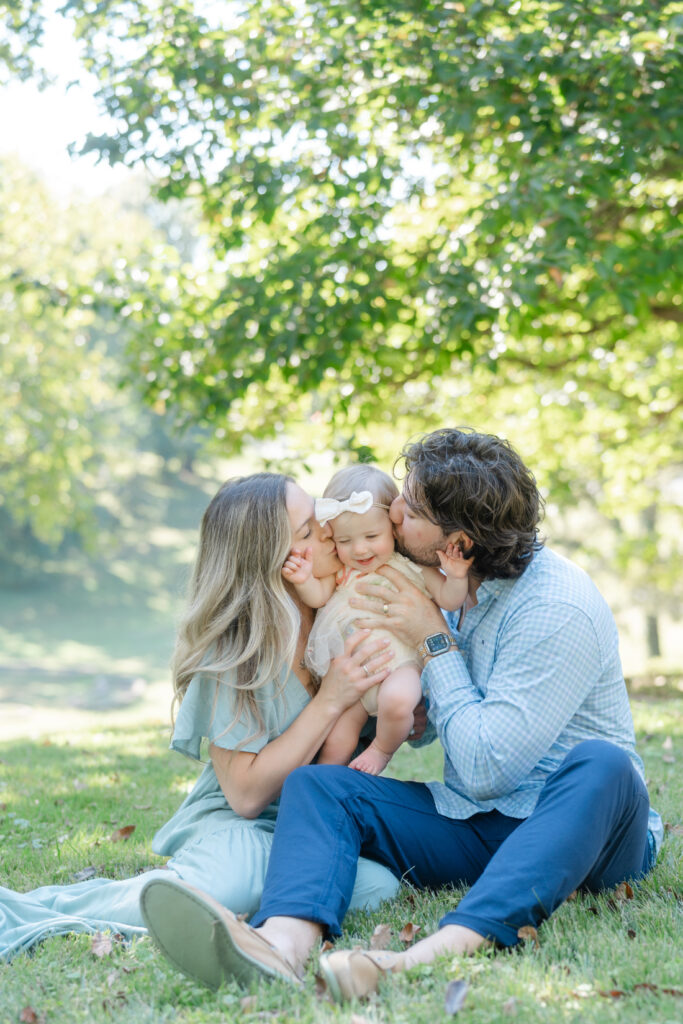 Nashville baby photography family moment of parents sitting in the grass, kissing their smiling one-year-old daughter.