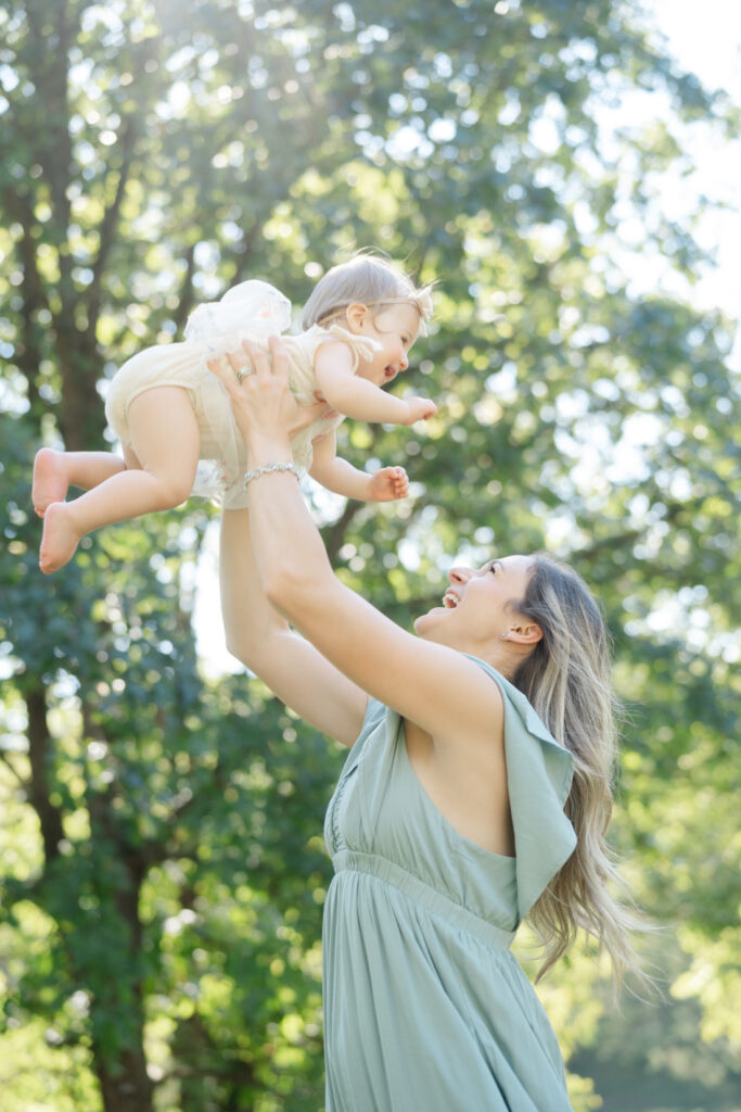 Nashville baby photography image of a joyful mother lifting her laughing one-year-old into the air beneath glowing sunlight.