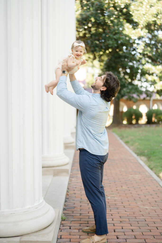 Father lifting his daughter into the air outside near tall columns as she laughs joyfully.