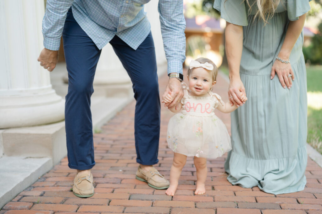 Nashville baby photography image of a barefoot one-year-old holding her parents’ hands while standing on a brick path.