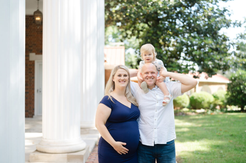 A family standing outdoors with their toddler riding on the father’s shoulders beside large white columns.