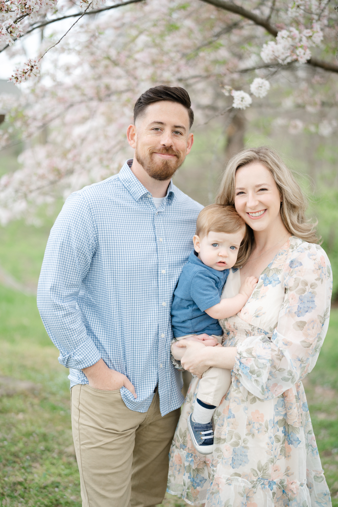 A family of three posing under blooming spring trees during their spring family photos session, with the parents holding their young child.