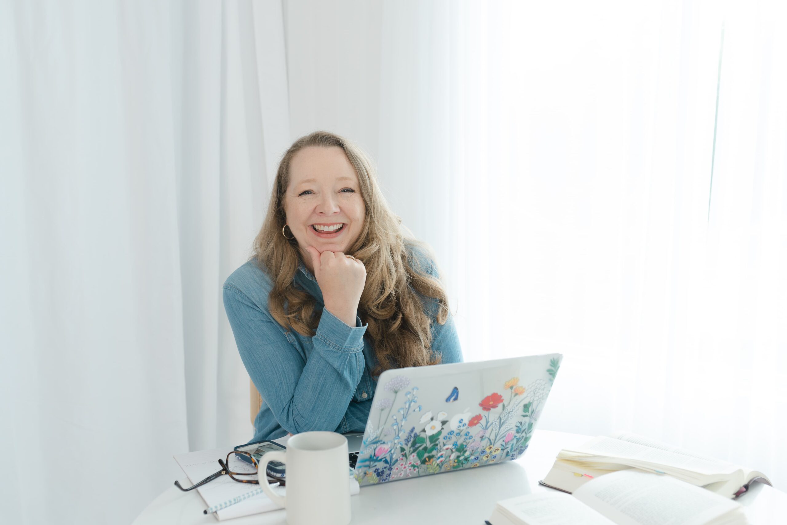 Woman seated at a white table with a laptop, books, and coffee mug, smiling thoughtfully in a bright studio environment.