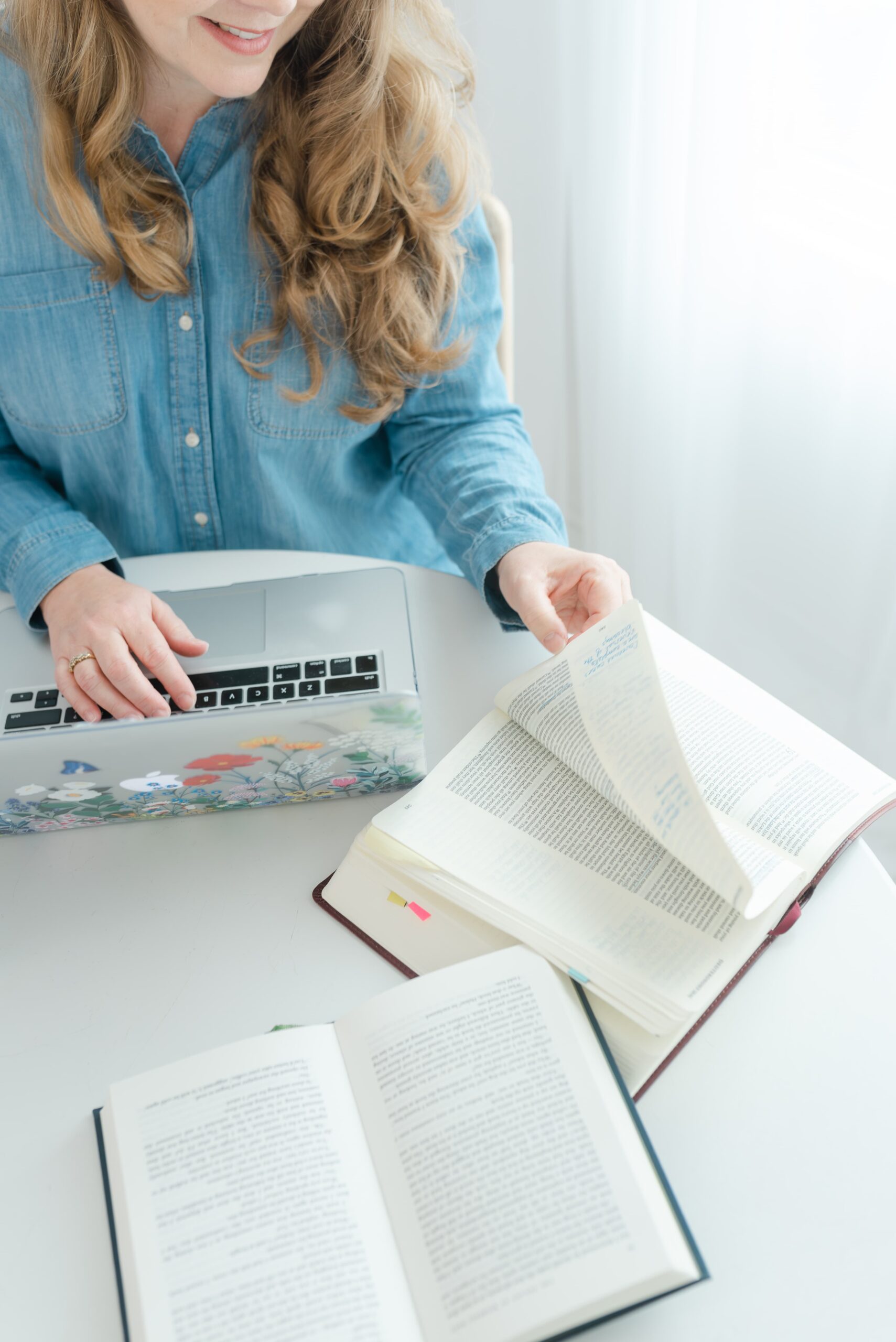 Woman flipping through an open book while typing on a laptop at a white table in a light-filled studio.