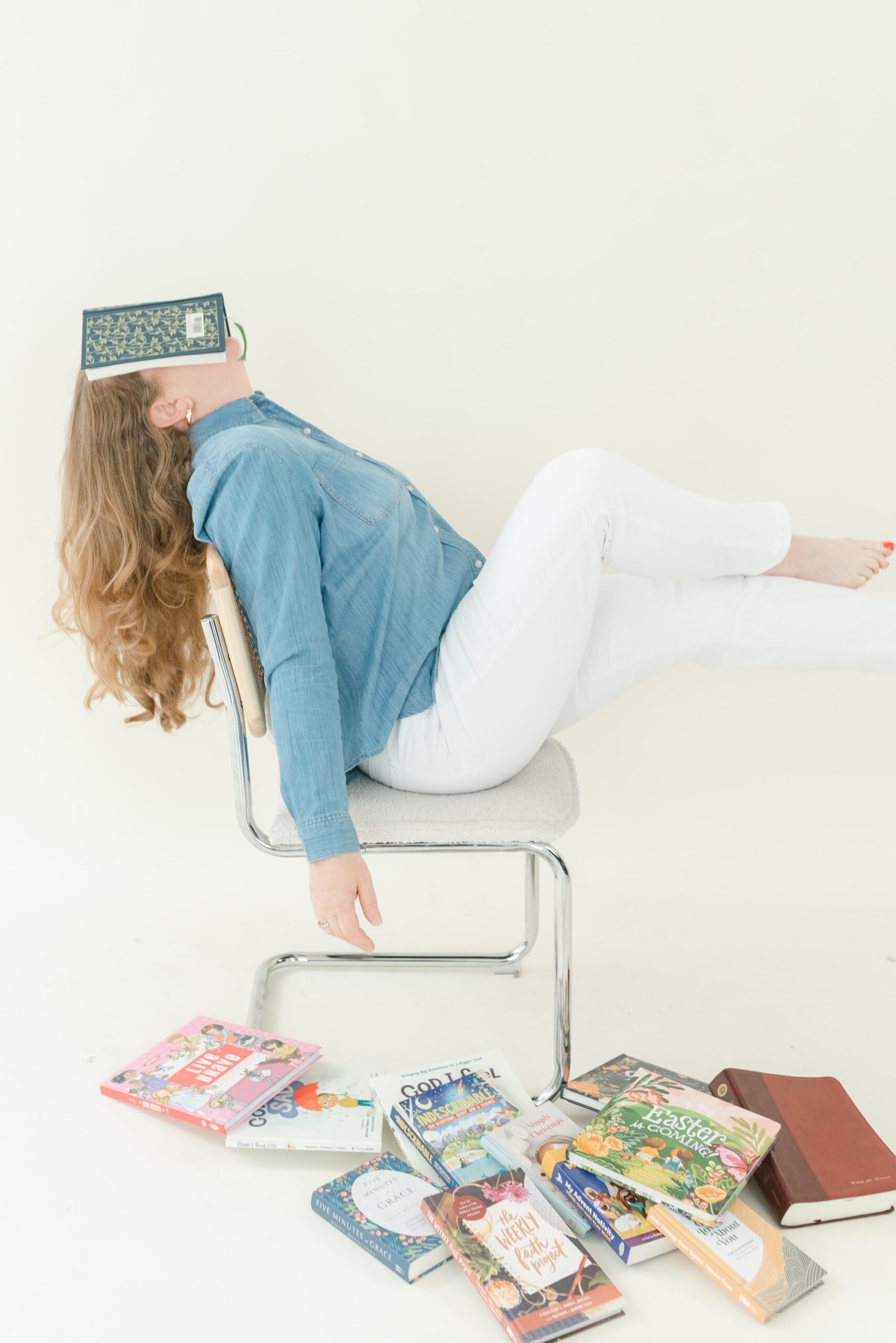 Woman reclining on a chair with a book resting over her face, surrounded by a stack of books on the studio floor.
