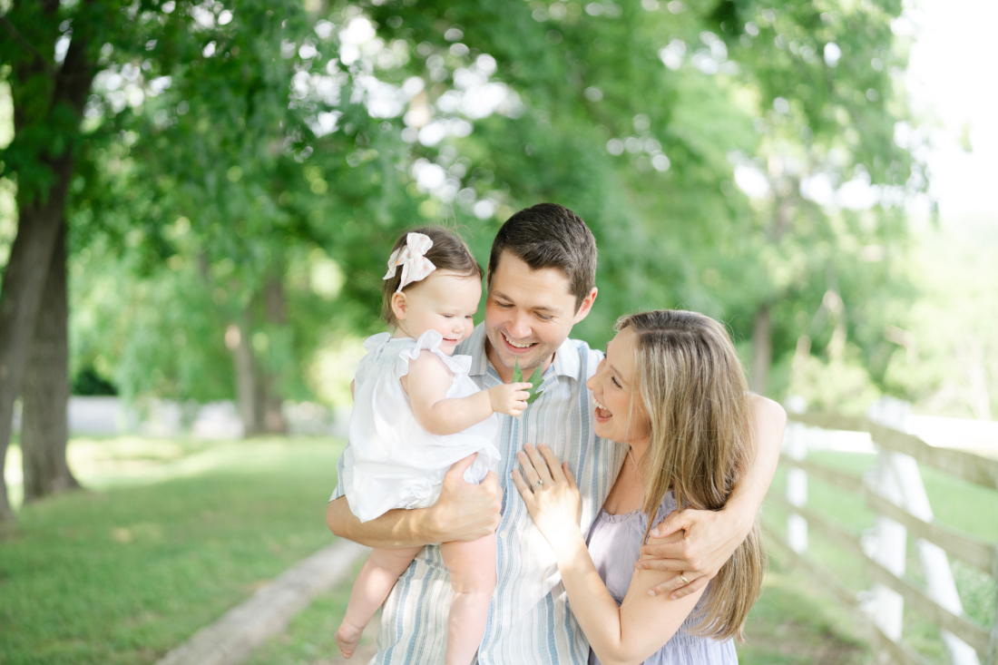 Outdoor family portrait of parents holding their baby while walking together on a green path, showing connection and movement naturally.