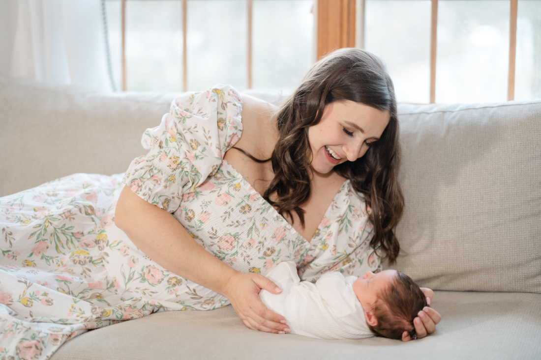Mother in floral dress holding newborn on couch during newborn photography session